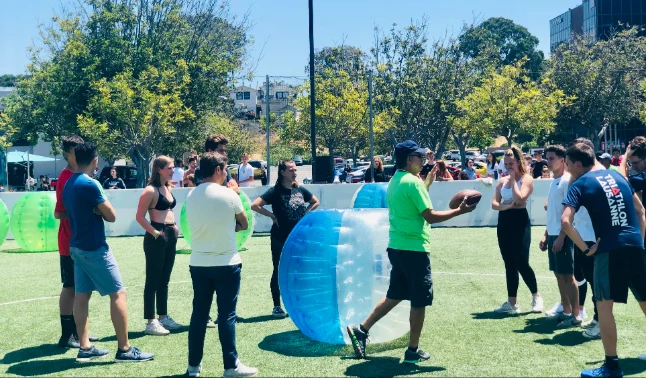Participants gathered on a soccer field, preparing for a bubble soccer game, with an instructor giving directions.
