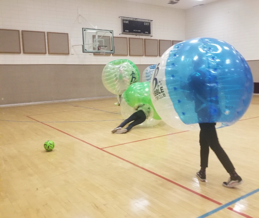 Participants in bubble soccer suits playing a game indoors, with one player on the floor and a soccer ball in view