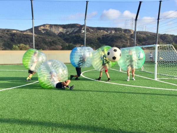 Participants in bubble soccer suits playing outdoors on a soccer field, with a large soccer ball in the air and one player on the ground