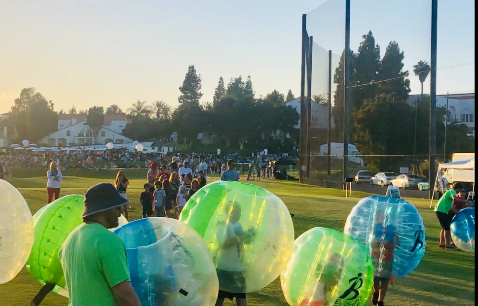 Participants in bubble soccer suits playing on a grassy field during an outdoor event, with a crowd of spectators and a sunset in the background