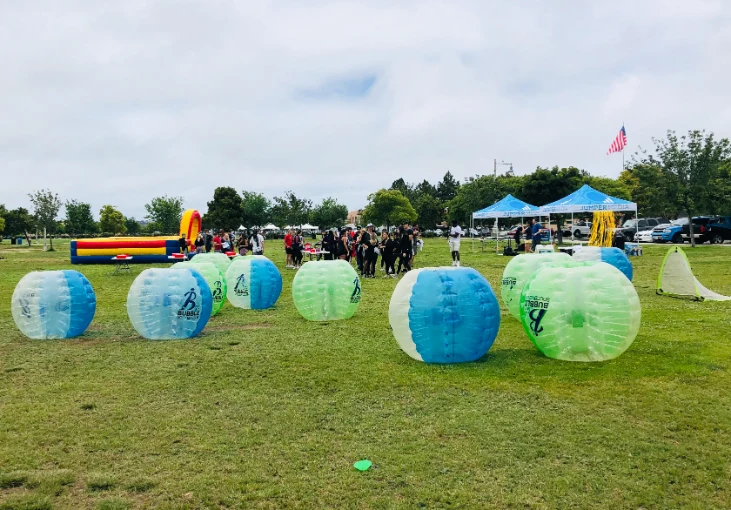 Outdoor bubble soccer event with multiple bubble soccer suits on the grass, participants in the background, and tents set up for the event